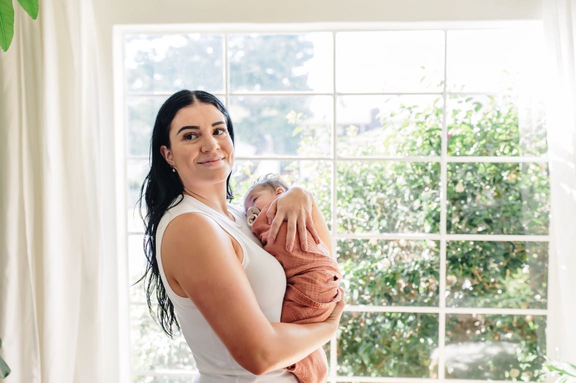 Ashlee standing in a sunlit room holding a sleeping baby in a rust-colored swaddle, smiling softly at the camera