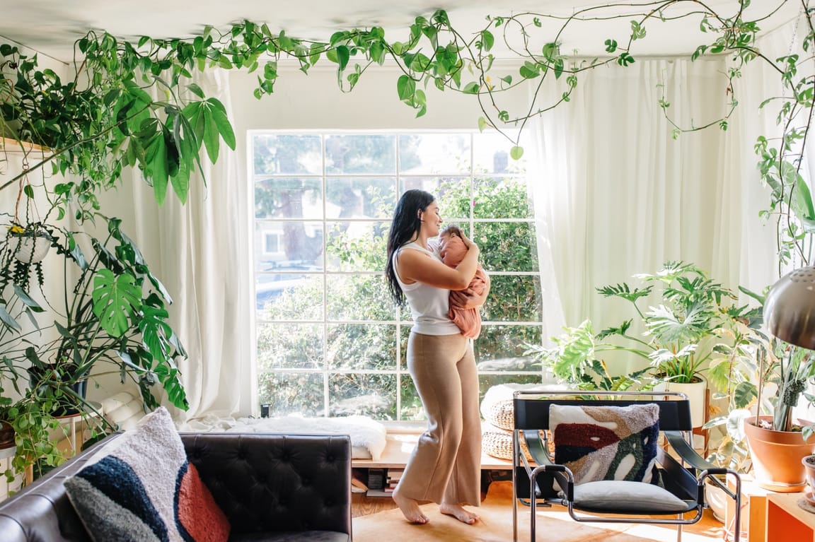 Ashlee standing barefoot by a sunlit window in a plant-filled room, holding a sleeping swaddled baby in profile