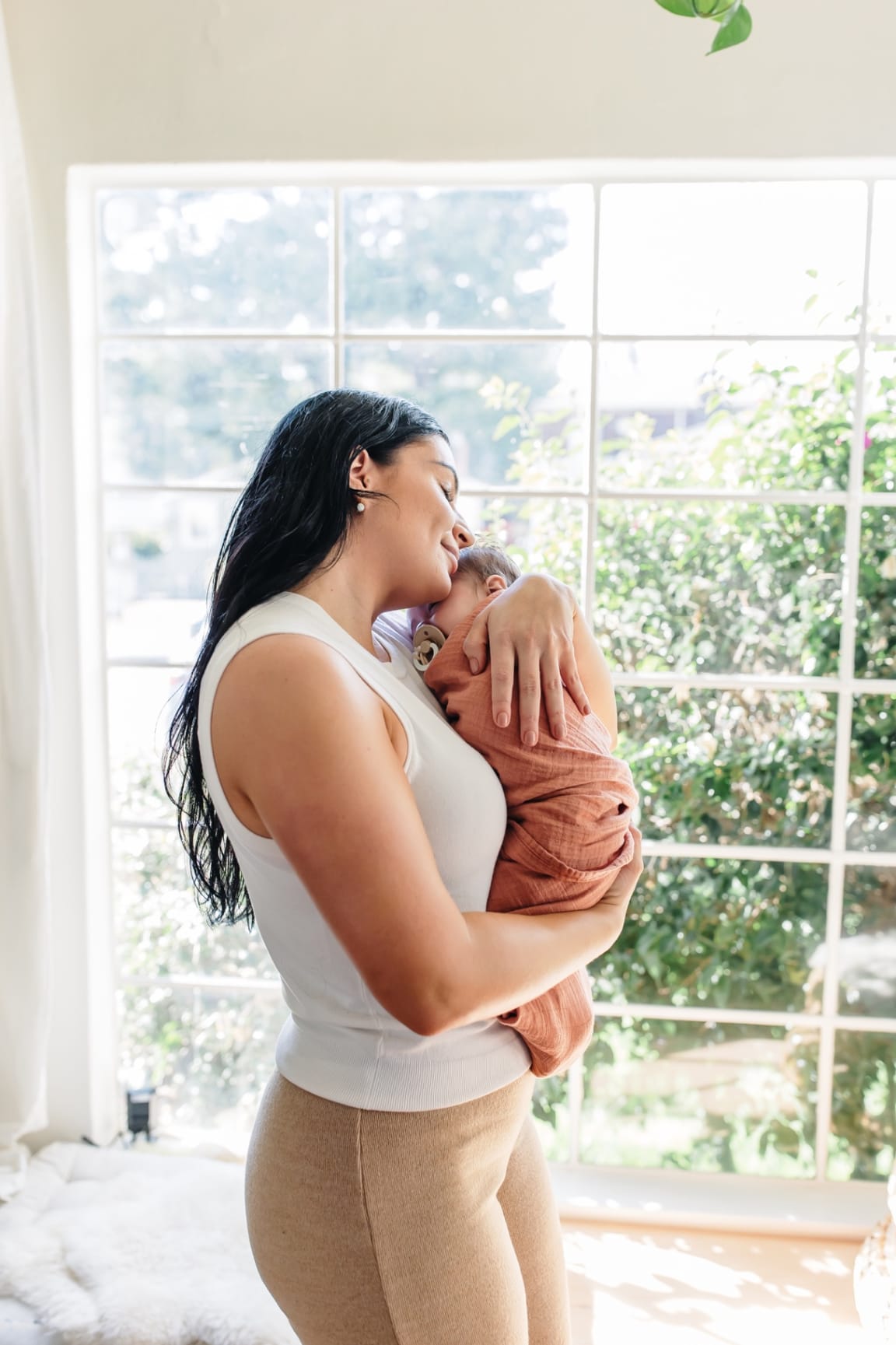 Ashlee cradling a swaddled newborn close to her chest in soft window light — a moment of attuned, tender presence
