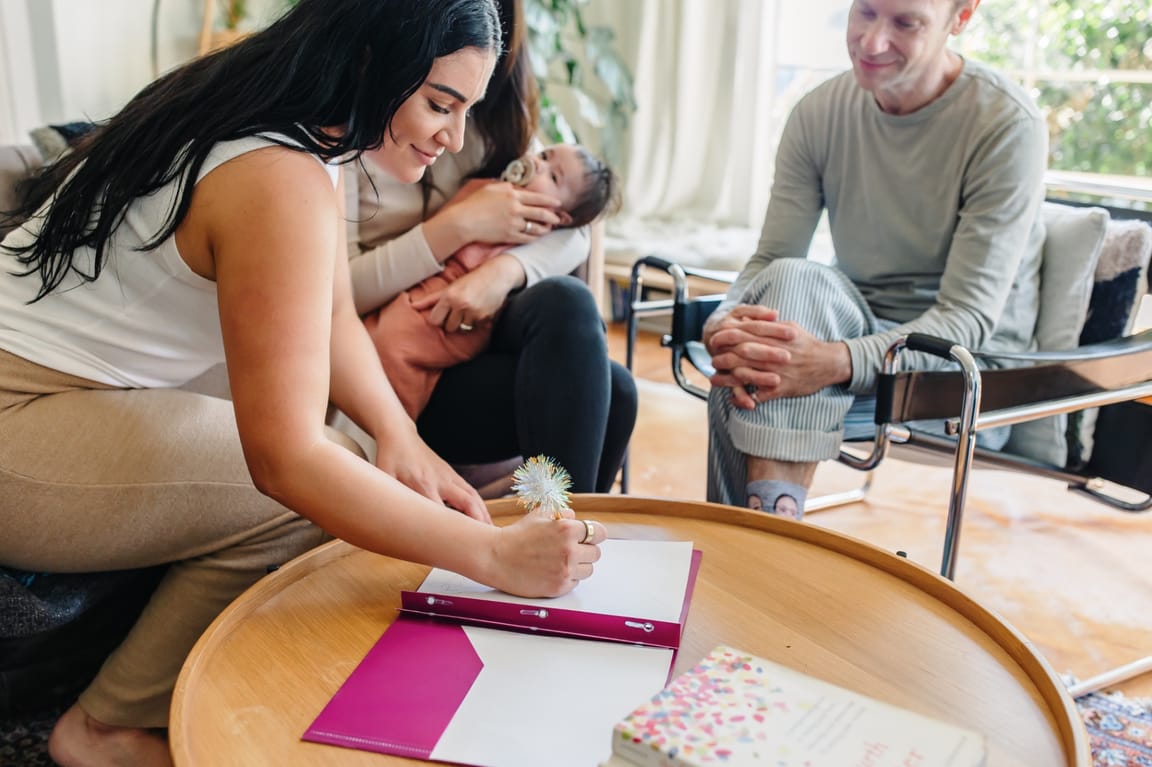 Ashlee taking postpartum planning notes at a coffee table while a parent gently holds their newborn nearby