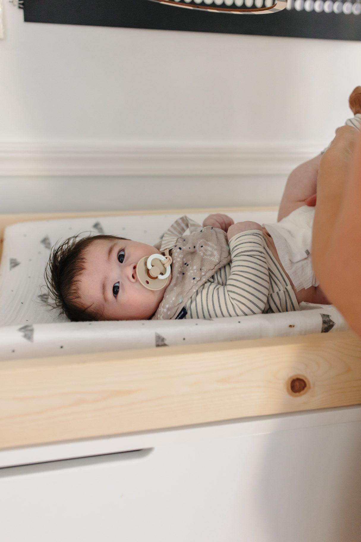 A wide-eyed baby with a pacifier resting on a changing table during a calm diaper change